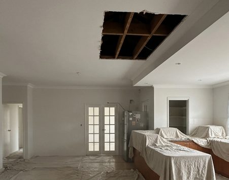 A section of ceiling taken out to the joists in an occupied house, with the furniture sheeted over below it.